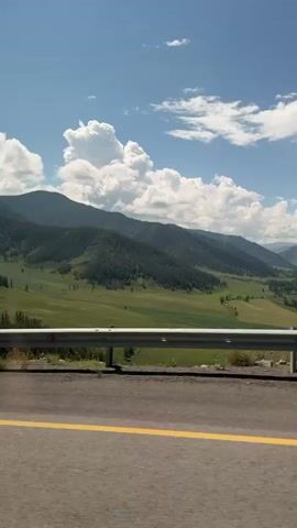This is a vertical video shot from a moving vehicle, looking out at a mountainous landscape. The road in the foreground is asphalt with a yellow marking. A metal guardrail runs along the edge of the road. Beyond the guardrail, the terrain slopes downwards into a valley filled with green fields and patches of trees. In the distance, several mountain ranges are visible, covered in dark green forests. The sky is blue with fluffy white clouds. The video pans slightly to the right, revealing more of the valley and mountains. No people or significant objects are clearly visible. The overall impression is of a scenic drive through a natural, rural environment.