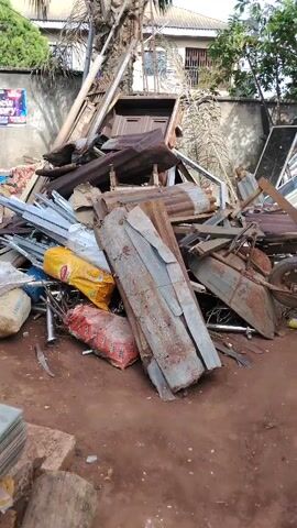 The video captures an outdoor scene in Akwa Etiti, Nigeria, dominated by a large, jumbled pile of scrap materials and discarded items. No people are visible.

The central focus is a mound of various materials including rusty corrugated metal sheets, wooden planks, metal frames (possibly from doors or windows), and other miscellaneous metal scraps. Amidst this pile, there's a small, rusty cart with a wheel. Several bags are also scattered around: a yellow bag with "DELICIOUS" partially visible, a red bag clearly labeled "RICE," and a white/beige bag where "CEMENT" can be seen.

In the foreground, to the left, a stack of what appears to be glass or clear plastic sheets rests on the ground next to a wooden log. The ground is reddish-brown dirt.

The background features a concrete wall, some green trees, and parts of a building with a metal gate or window. A blurry poster is visible on the wall in the distance. The scene suggests a storage area for construction debris or recyclable materials. The lighting indicates it is daytime.