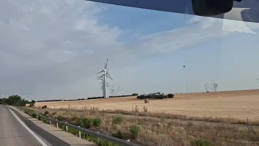 A video taken from a moving vehicle shows a rural landscape dominated by a wind farm. In the foreground, a paved road curves to the left, bordered by a metal guardrail and dry vegetation. Beyond the road, a vast, golden field stretches out, appearing to be recently harvested. A single, large, cylindrical bale of hay sits in the middle of this field. A line of tall wind turbines stands prominently in the midground, stretching from left to right across the frame. These turbines have white towers and three angled blades. Further in the distance, more wind turbines are visible, scattered across the horizon. The sky above is partly cloudy with a mix of blue sky and white clouds. A dark, small object, possibly a bird or a drone, is visible against the sky in the upper right quadrant of the frame. The overall impression is one of open, agricultural land with a significant presence of renewable energy infrastructure.