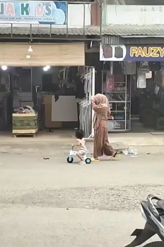The video shows a street scene in Keudegeudong, Indonesia. The primary subjects are a young child riding a small toy bicycle on the street and an adult woman wearing a hijab and striped attire, who appears to be watching the child or taking a photo. In the background, there are several storefronts. One prominent storefront has a sign that reads "Nanja Kids" in blue and white letters, with smaller text below it. This shop appears to sell children's clothing and accessories, as indicated by racks of clothing visible inside. Another shop to the right displays various items on shelves, including what look like backpacks.

The activity taking place is seemingly mundane, with the child enjoying a ride on their toy while the adult supervises. Two people on a red scooter ride past in the background, adding to the sense of everyday life on the street. The time of day appears to be daytime, with natural light illuminating the scene. The weather seems fair. The setting is a street in front of shops, suggesting a commercial area. The overall mood is relaxed and observational, capturing a slice of local life.