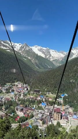 The video shows a view from a cable car ascending a mountain. In the foreground, the dark cables of the cable car are visible, and the edge of the cable car window. Below, a small town with colorful buildings is nestled in a valley surrounded by lush green forests. In the background, majestic snow-capped mountains rise against a clear blue sky. The camera pans slightly, offering different perspectives of the town and the mountain range. There are no people clearly visible in the video, only the town and the natural landscape. The lighting suggests it is daytime, likely with bright sunshine.