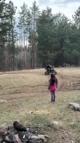 The video shows a girl standing in a field with trees and a fire pit in the foreground, looking at two all-terrain vehicles (ATVs) in the distance. One ATV is small and yellow with two people on it. The other is larger, black and white, with a roll cage. The girl runs a few steps toward the camera, then stops and watches as the black and white ATV drives closer. The ATV makes a sharp turn, kicking up dirt. The girl is wearing a pink shirt, a patterned jacket, and leggings. The sky is overcast.