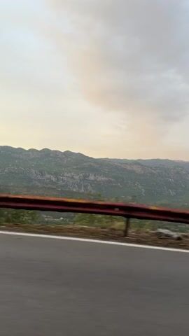 The video shows a moving car on a road with a guardrail. The mountains are visible in the distance, with trees on the side of the road. The sky is cloudy and overcast.