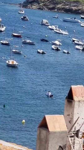 This is a high-angle video of a body of water filled with many boats of various sizes. In the foreground, there are two rooftops with visible chimneys. The water is a deep blue, and the sky appears clear. A single person is visible in the middle of the water, paddling a small boat, possibly a kayak or paddleboard. The boats are anchored and appear to be in a bay or harbor. The coastline with rocky terrain is visible in the background. The lighting suggests it is daytime.