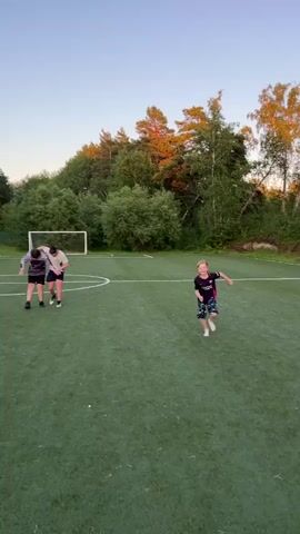 Two young boys are on a grassy field, possibly a soccer field, based on the presence of a goal in the background. One boy in the foreground, wearing a dark t-shirt and patterned shorts, is running towards the camera with a smile. Further back on the field, another boy in a black t-shirt and shorts is playing with a third boy who is bent over. The sun appears to be setting, casting a warm glow on the trees and sky. The overall scene depicts children playing outdoors during the evening.