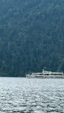 A white passenger ship sails on a large body of water. The ship has multiple decks and windows, and a Russian flag is visible on its roof. The ship is positioned on the left side of the frame. In the background, a densely forested mountain slopes down to the water's edge. The water's surface is choppy, with small waves. The sky above the mountains is partly cloudy. The camera is moving, as indicated by the slight motion blur and the water splashing at the bottom of the frame in the later part of the video.