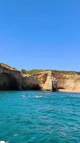 The video shows a bright, sunny day with a clear blue sky over a vibrant turquoise ocean. In the foreground, the water is choppy with whitecaps. In the midground, a rocky cliff face dominates the scene, with several caves carved into its side. Some of these caves appear to be accessible by boat, with small boats visible near the openings. There are also several people in the water, some on paddleboards and one in a kayak. In the background, a sailboat with a single sail glides across the water. The overall impression is one of a beautiful, active coastal environment on a summer day.