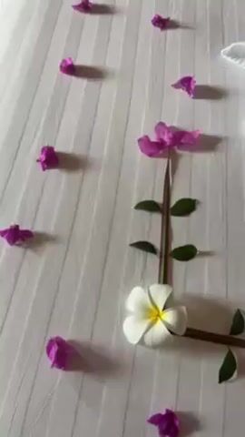 The video shows a decorated bed with white sheets. Pink flower petals are scattered on the bed. There is a white flower with a yellow center near the corner of the bed. Green plant stems and leaves are arranged to form a flower stem. There are two white towels folded into the shape of swans facing each other. The towels have white flowers with yellow centers placed in the folds. The words "HAPPY HONEY" are spelled out using green plant stems.