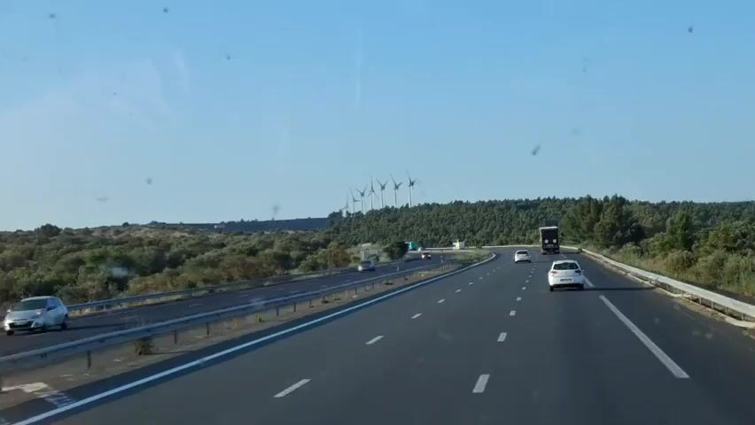 A highway scene unfolds under a clear blue sky. A multi-lane road curves gently to the right, with white lines marking the lanes. Several vehicles are visible on the road, including cars and a truck. On the left side of the highway, a gentle slope is covered with sparse vegetation and some scattered trees. Further in the distance, a line of wind turbines stands on a hilltop, silhouetted against the sky. The landscape is arid, with dry brush and scattered trees. The overall impression is a sunny, rural highway in a wind farm region.