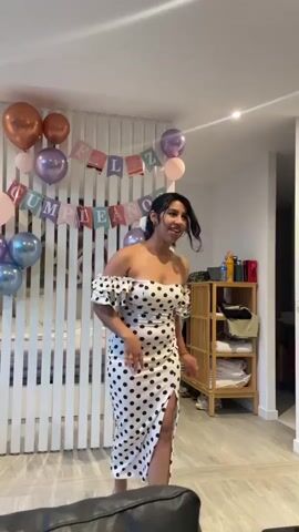 A young woman is dancing in front of a white slatted wall. She is wearing a white off-the-shoulder dress with black polka dots and a high slit on the left side. The dress has short, ruffled sleeves. Above her, a banner reads "FELIZ CUMPLEANOS" and is decorated with balloons in shades of pink, purple, and copper. To her right, there is a wooden shelving unit with various items on it, including bottles and what appear to be rolled-up towels or blankets. The lighting is warm and appears to be coming from overhead fixtures. The floor is a light-colored wood. The overall impression is a festive home environment, possibly a birthday celebration. The woman is smiling and appears to be enjoying herself. Her hair is dark and pulled back. Her arms are in motion as she dances.