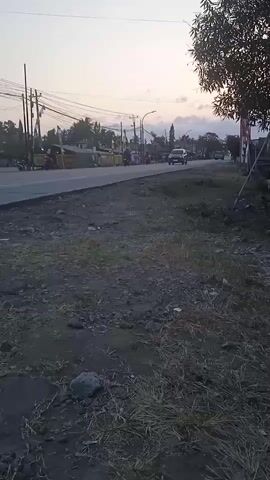 The video shows a road with several vehicles. The sky is overcast with a hint of sunset. There are trees and power lines on the side of the road. The ground is covered in dirt and grass. The video appears to be taken from a low angle. There is a slight tremor to the camera as if it is being held.