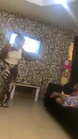 A woman is dancing in front of a TV in a living room, while a toddler is lying down on a couch. The woman is wearing a white tank top and patterned pants. She's facing the TV and making dance movements with her hands and body. The TV is mounted on a wall with a swirling black and white patterned wallpaper.  A small, light-colored table stands near the TV. To the right, a black couch is visible, and a child in a pink shirt lies on it, partially covered by a colorful blanket. Another person is sitting on the couch with their legs visible. To the right of the couch there are children's items, including a colorful box with shapes. The room is well-lit, and there is a skylight or ceiling light visible.  The music is playing loudly.