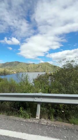 The video shows a motorcycle ride, with a scenic view of a lake and surrounding hills. Initially, the camera is positioned looking past a guardrail at a body of water surrounded by green hills under a partly cloudy blue sky.  The road is seen curving upwards along the hillside. Then the view switches to looking down on two people riding on a motorcycle. The person in the front is wearing a black leather jacket with red accents and a white helmet. The person in the back is wearing a grey backpack.  The camera angle shifts slightly as the motorcycle travels down the road, and a white truck passes in the opposite direction.