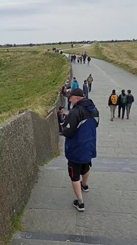 The video shows a man in a blue jacket and black shorts standing by a stone wall, looking at his phone. He is on a paved pathway next to a grassy hill. Several other people are walking on the pathway, some in the distance. Further down the path, a woman is sitting by the wall playing a flute. In the far distance, a tower is visible on the horizon. The sky is overcast.