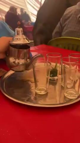 The video shows someone pouring tea from a silver teapot into small glasses on a silver tray.  The tray sits on a red tabletop inside a large tent, which is filled with people sitting at tables.  There are several other glasses on the tray, one of which contains fresh mint.  Steam rises from the tea and the tent appears crowded.  The interior of the tent looks worn and the roof has many seams.