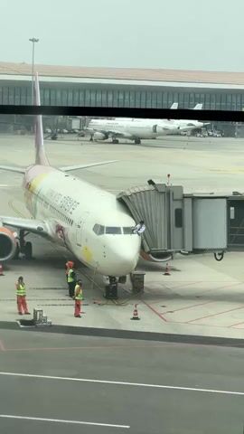 The video shows an airplane parked at the airport with a walkway attached. The airplane is white with orange accents, with some colorful cartoon graphics on the body. The text "CHINA UNITED AIRLINES" is printed on the side of the plane. Several ground personnel are working near the plane wearing bright orange vests. In the background, another airplane, labeled "CHINA EASTERN", can be seen parked further away. The video appears to have been taken through a window.