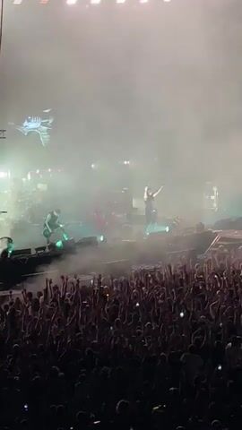 The video shows a concert scene. In the foreground, a large, dense crowd of people is visible, with their arms raised in the air, facing a stage. The crowd is mostly dark silhouettes. On the stage, there are several musicians performing. One musician, center-stage, is singing or speaking with an arm raised. Another musician, to the left, is playing a guitar. Behind them, drums and other stage equipment are visible. The stage is filled with smoke or fog, and bright lights are illuminating the performers. A large screen or projection is visible in the upper left of the stage, displaying a graphic. The overall atmosphere appears energetic and celebratory.