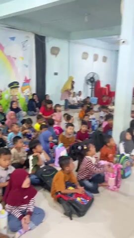 A group of children and adults are gathered in a room. The children are sitting on the floor, many with backpacks in front of them. Some of the adults are standing, and some are also sitting on the floor. There is a colorful mural on the wall behind the children, featuring a rainbow, cartoon characters, and butterflies. Red chairs are stacked in the background. The room appears to be brightly lit.