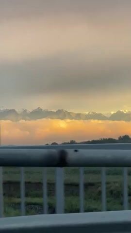 The video shows a view of a mountain range partially obscured by clouds. The sun is setting or rising, casting an orange glow on the clouds. The mountains appear to be snow-capped. In the foreground, there is a metal railing and a hint of green foliage, suggesting the view is from a bridge or elevated walkway. The overall atmosphere is peaceful and scenic.