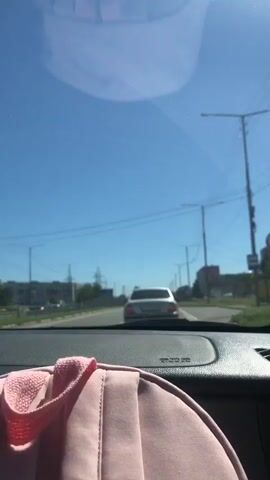 The video shows a view from inside a car as it travels down a road on a clear, sunny day. The camera is looking forward through the windshield. In front of the car, another white car is also driving down the road. The road is paved and has multiple lanes. On the left side of the road, there is a fence and some buildings in the distance. On the right side, there are more buildings and utility poles with wires. The sky is a bright blue and a reflection of something, possibly the car's interior, is visible on the windshield. The dashboard of the car is partially visible in the foreground, with a pink cloth item on it.