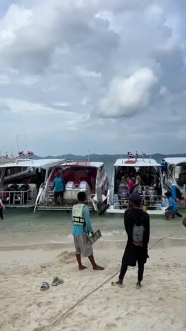 A man wearing a life vest stands on a sandy beach, holding a clipboard. He is facing away from the camera. To his right, a person in a black outfit with a backpack stands looking towards boats. Several boats are docked in the clear water, some with people on board. The sky is cloudy. Shoes are on the beach near the man.