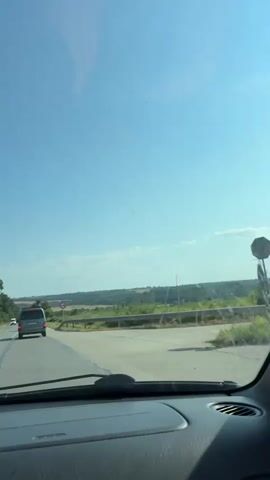 The video, filmed from inside a vehicle, shows a clear blue sky with a few wispy clouds. The view is through the windshield, which has a faint reflection on its surface. In the distance, rolling hills covered in greenery are visible, suggesting a rural or semi-rural landscape. A road runs through the scene, and a silver minivan is traveling ahead on the right lane. A round red and white traffic sign, indicating "No Entry" or "Do Not Enter," is positioned on the side of the road to the right of the minivan. Guardrails line the side of the road. The vehicle appears to be moving along a highway or a well-maintained road. The time stamp indicates the video is very short, with no significant events occurring beyond the visual depiction of the drive.