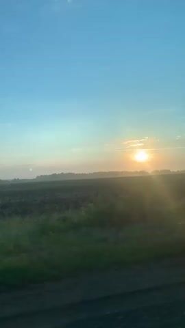 The video shows a view from a moving vehicle through a window. The scene is a rural landscape with a field and trees. The sun is low on the horizon, suggesting sunrise or sunset, and is partially obscured by trees. The sky is a clear blue with some wispy clouds. The ground in the foreground appears to be a dirt road. The overall impression is one of a peaceful, natural setting.