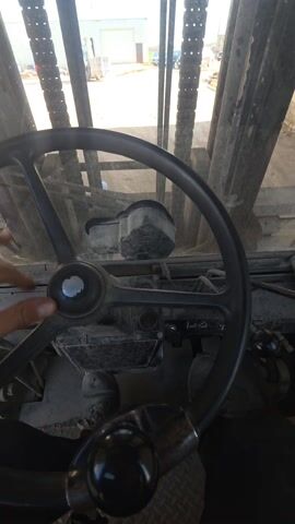 The video shows the inside of a piece of heavy machinery, probably a forklift or similar vehicle, with the view looking out through the metal forks. A hand is visible, gripping a handle near the steering wheel.  Outside, there's a dusty yard with a building and other vehicles, including a red forklift in the distance. The sky is blue. The inside of the vehicle looks dirty and well-used.
