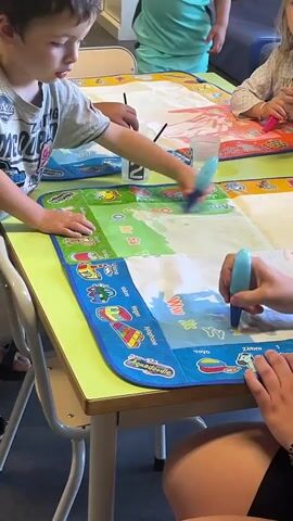 This is a close-up shot of children and an adult sitting around a table, engaged in a coloring activity. The table is covered with a large, colorful mat, divided into sections with illustrations and text, possibly educational in nature. Several children are holding water-filled paint pens and drawing on the mat. The pens are blue and have a brush-like tip. One child, a boy wearing a grey t-shirt, is seen looking down at the mat with a concentrated expression. Another child's hands, likely an adult's due to the presence of a bracelet and tattoos, are visible holding a pen and drawing on the mat. In the background, other children are also participating in the activity. The environment appears to be a classroom or a play area, with other tables and chairs visible. The lighting is bright and even, suggesting an indoor setting.
