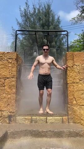A man in black shorts and sunglasses stands at the bottom of a stone stairway between two stone walls, as water falls from a black metal bar at the top of the steps. He walks up the steps, placing his hands on the walls as he goes, and stops midway up, looking into the camera. He has a slight smile on his face and seems to be enjoying the water. He continues to walk up the remaining steps.  Behind the stairway is a tree and a bright blue sky with a few clouds.