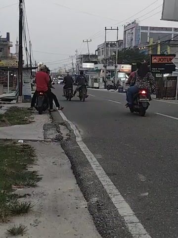 The video captures a street scene at dusk. A few motorcycles are visible on the road. One motorcycle in the foreground has two people on it, the driver wearing a red shirt and the passenger wearing a dark shirt. The motorcycle is moving away from the viewer and slightly to the right. Other motorcycles are further down the road, also moving in the same direction. The environment appears to be a town or city with buildings and streetlights lining the road. A sidewalk is present on the left side of the road with some vegetation. The sky is overcast.