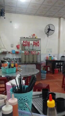 The video shows the interior of a casual restaurant in Vietnam. The restaurant has a simple tiled floor, white walls, and a tiled ceiling with fluorescent lighting. Several ceiling fans are visible. There are multiple metal tables with red plastic stools. A glass display case labeled "COM-PHO HANOI 3" is present near the entrance, filled with various food items. Another glass case, labeled "TRUNG" and containing what appears to be eggs or other goods, is near a "Tra cascara" advertisement poster. Several people are seated at tables, some wearing masks. One man is sitting on a stool, appearing to be talking on a phone. The tables are set with chopsticks in a blue holder, small black bowls, and condiment bottles. The overall impression is a typical, unpretentious Vietnamese eatery.