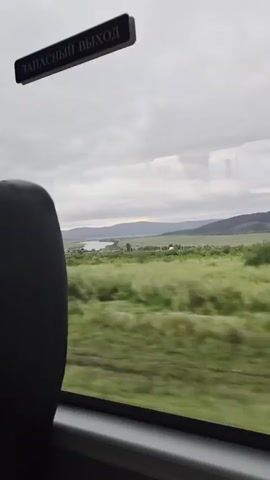 A blurry shot from inside a vehicle shows a sign reading "EMERGENCY EXIT" in Russian. Outside the window, a landscape of green fields and hills stretches to the horizon under a cloudy sky.