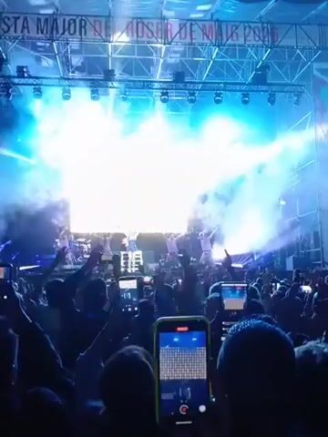 The video depicts a live concert with a large crowd recording the performance on their phones. The stage is illuminated by bright lights and filled with smoke, partially obscuring the performers, a lead singer in a blue outfit with dancers dressed in purple. The audience is packed tightly together, with numerous phones raised to capture the event, creating a sea of screens. A banner with text on it is visible towards the upper right corner of the frame. The overall atmosphere is lively and energetic.