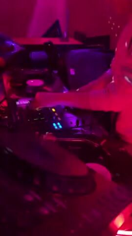 In a dimly lit room with red and purple lighting, a person operates a DJ setup. The equipment includes two turntables with records spinning, a mixer with lit buttons, and a microphone stand nearby. Speakers are visible in the background. The person wears a light-colored top and manipulates the mixer's controls with their hand. The scene suggests a DJ performing or mixing music in a club or studio setting.