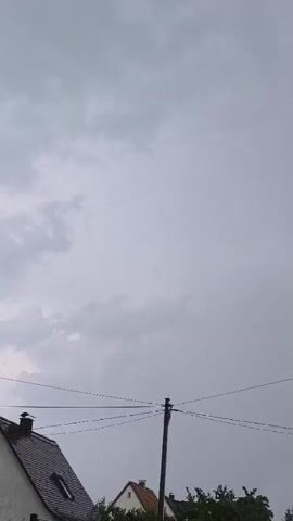 The video shows a view of the sky on a cloudy day. There are some faint vertical lines visible in the sky, which appear to be rain. In the foreground, the roof of a house with a chimney and a pitched roof is visible on the left. A utility pole with wires is in the center of the frame. Trees are visible on the right. The overall impression is of a grey, overcast day with light rain.