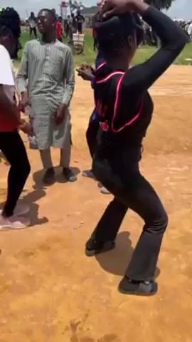 A group of three young women, one wearing a black hijab, dance together outside on a dirt lot in broad daylight, surrounded by other people standing and watching. The woman in the middle wears a white t-shirt and the woman on the right wears all black with a sports jersey. A young man wearing a light-colored, patterned robe-like shirt stands slightly behind them, swaying slightly to the music. In the background, other people stand and watch, some under a white parasol or other forms of shade. The sky is partly cloudy.