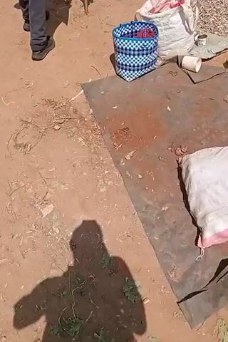 The video shows an outdoor market scene. In the foreground, a shadow of a person is cast on the dirt ground. To the right, a blue and white woven basket is visible, containing some items. A large white sack with pink stripes lies in the lower right corner. In the middle ground, several buckets filled with potatoes are arranged on the ground. A few people are walking in the background. One person is wearing a light blue shirt and dark pants, another is wearing a brown t-shirt and blue jeans, and a third person is wearing a dark shirt and pants. The ground is a mix of dirt and some scattered debris. There are also some market stalls and structures visible in the background.