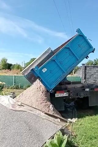 A blue dump truck is parked on a grassy area with a gravel pile in the foreground. The truck's bed is tilted upwards, and sand is being dumped from it onto the gravel. The sky is clear and blue.