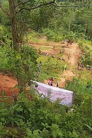 A person wearing a red and black outfit is riding a dirt bike on a narrow dirt track through a dense forest. The rider loses control and falls off the bike, which lands on its side. The rider appears to be getting up. The scene is filmed from a high angle, looking down on the track. There are ropes running along the side of the track.