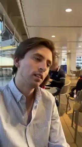 A person with short brown hair and a light blue striped shirt is talking to the camera. The person says "Emily, happy birthday, I love you a lot." The person is indoors. In the background are tables and chairs and other people, along with windows.