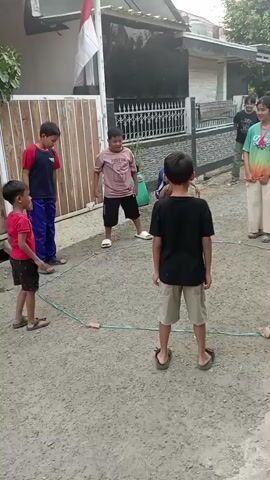 The video shows a group of children playing a game in a dirt yard. A blue rope is laid out in a hexagonal shape on the ground. Six children are visible, five boys and one girl. One boy in a red shirt and black shorts stands to the left, facing inwards towards the center of the hexagon. A boy in a blue shirt and dark pants stands behind him, also facing inwards. Another boy in a light pink shirt and black shorts stands in the upper center of the hexagon, looking towards the center. A boy in a black shirt and tan shorts stands in the lower right of the hexagon, facing away from the camera and towards the center. A boy in a green and tie-dye shirt and white pants stands to the right of the hexagon, facing inwards. A girl in a black and white patterned shirt and black pants stands further to the right, also facing inwards. In the center of the hexagon, a boy in a black shirt and tan shorts is in mid-air, jumping over something on the ground. Two small brown objects, possibly stones or small toys, are placed at two points along the rope. A red and white flag is visible on a pole to the left, attached to a building. The environment appears to be a residential area, with houses and a fence in the background. The children are interacting with each other as part of the game.