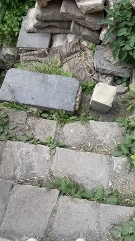 The video begins with a close-up view of large, uneven grey stones and some green vegetation. A person's legs, wearing white sneakers and blue jeans, are visible in the foreground, stepping on the stones. The camera then pans up and to the right, revealing a pile of stacked stones and a patterned fabric underneath. More green plants are growing amongst the stones and on the ground. The scene appears to be outdoors, possibly in a historical or archaeological site. The latter part of the video shows a long, stone wall with battlements, likely part of a castle or fortress. The wall is made of grey stones and has a walkway on top where a few people can be seen. The surrounding environment includes more greenery and a distant view of mountains under a cloudy sky. The overall impression is one of an old, weathered structure integrated with nature.