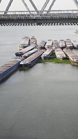 The video begins with an overhead shot of a river, with a bridge spanning across it. Several barges are moored together in the water, clustered near a small island or sandbar. The barges are long and flat-topped, appearing to be loaded with cargo. The bridge is a large, metal structure with an intricate lattice design. As the camera pans, it shows the stone pylons of the bridge supporting the structure. Further down the river, another barge is visible. The camera then continues to pan along the river, showing the banks lined with trees and a red buoy floating in the water. The sky is overcast, and the overall lighting suggests it is daytime.