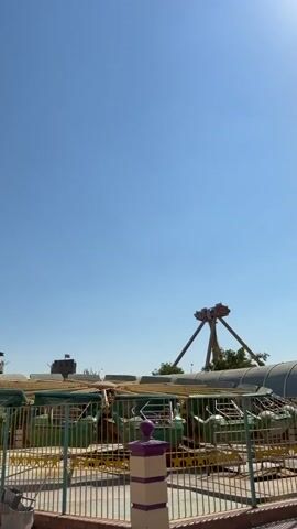 The video shows an amusement park ride with a circular platform with multiple gondolas or seats. The ride is in motion, with the platform spinning and the gondolas tilting. In the background, there is a larger ride with two large arms that pivot. The sky is clear and blue, indicating a sunny day. There are some fences and railings around the ride. Two people are visible standing near the fence, facing away from the camera and towards the ride.