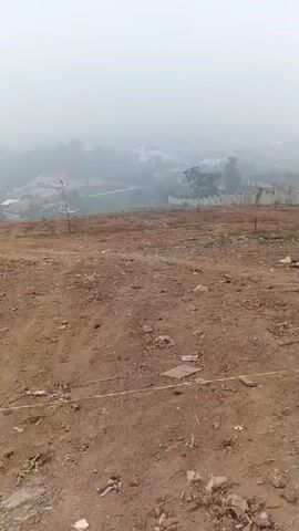 The video shows an outdoor scene from a slightly elevated perspective, looking down a gentle slope. The ground in the foreground is rough, unpaved earth with some scattered debris, including what appears to be plastic packaging and a blue cylindrical object. A yellow marking tape runs horizontally across the lower part of the frame. In the middle ground, there is a single red and white flag on a pole. Beyond that, the landscape descends into a valley filled with trees and some buildings, obscured by a dense fog or mist. The sky is completely overcast with a light gray fog. There are no people visible in the video.