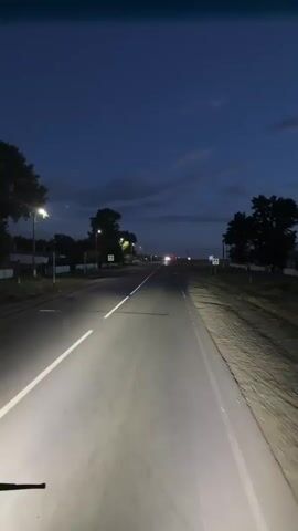 This is a first-person perspective video clip of a drive on a road at dusk, likely in Baiqadam, Kazakhstan. The scene is characterized by a dark, twilight sky with scattered clouds. The road is asphalt and well-maintained, with clear white lane markings. Headlights from the vehicle create bright beams illuminating the road ahead.

The environment is rural or semi-rural, with trees lining the sides of the road and some distant buildings visible. Streetlights are present, casting pools of light. There are also prominent road signs, including pedestrian crossing signs with a yellow triangular background and a black pedestrian silhouette. A bus stop sign is also visible on the right side of the road.

The primary activity is driving, moving forward along the road. The time of day is clearly transitioning from day to night, indicated by the deep blue of the sky and the artificial lighting. The overall mood is calm and perhaps a little introspective, fitting for a drive during twilight. There is no visible text in the video clip itself, other than the text on the road signs.
