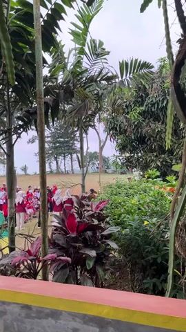 The video opens with a view through foliage and a structure, likely a covered patio or porch. In the background, a group of people, mostly young individuals dressed in red and white uniforms, are standing in an open grassy field. Some of the individuals are wearing headscarves. The group appears to be participating in an outdoor activity or ceremony. There is a tall, colorful, rocket-shaped object positioned on the field, leaning at an angle. The environment is outdoors with trees and vegetation visible. The sky is overcast. The video is brief and appears to capture a moment from a distance.