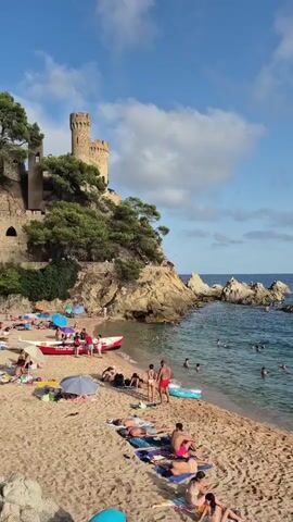 A bustling beach scene unfolds with a medieval castle perched atop a rocky cliff in the background. The sand is dotted with numerous people, many sunbathing on towels or laying on lounge chairs. A few individuals are visible in the turquoise water, some swimming and others relaxing on inflatable floats. Several colorful umbrellas are scattered across the beach, providing shade. A small red boat is pulled up on the shore near a cluster of people gathered together. The sky is a bright blue with scattered white clouds.
