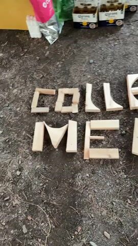 The video pans over an outdoor scene on the ground. In the foreground, wooden letters are arranged to spell out "COLLECT MEMORIES". The letters are various shapes and sizes, made of light-colored wood and appear to be handmade. The ground is covered in dirt, pine needles, and small stones. In the background, there are several cases of Corona beer on a pale surface, possibly a table or tarp, with a pink bag to the left of the beer cases. Some green foliage is visible in the upper background. The video is shot from a high angle, looking down at the scene.